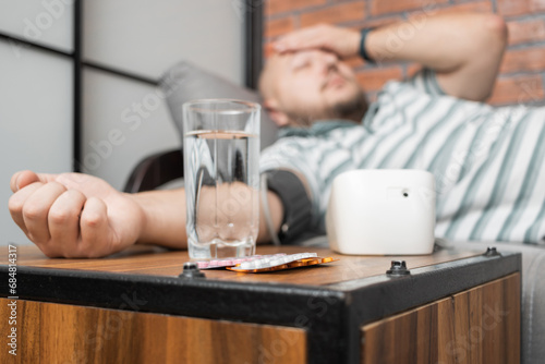 Defocused background with man who has hypertension and headache, lying on sofa at home, measures blood pressure on hand with modern digital tonometer, focus on blister pack with pills on foreground