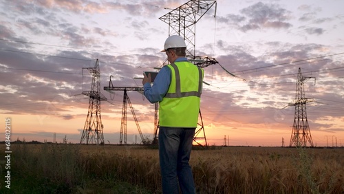 Electrician in helmet checks data on tablet walking to power transmission lines photo