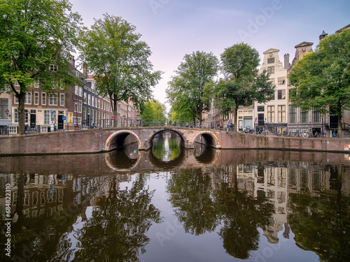 Arch bridge over the Herengracht and Leidsegracht canals in the historic center of Amsterdam on an early summer Sunday morning. Historic canal houses reflect in the calm water.  photo