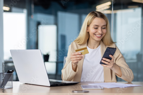 Young joyful woman at workplace inside office holds phone , bank credit debit card in hands, businesswoman chooses gifts in online store, book service and transfer funds using app on smartphone