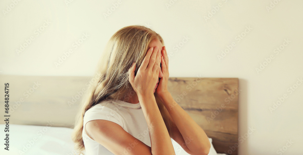 Upset sad crying middle-aged woman covering her eyes with her hands while experiencing mental or physical suffering sitting on bed at home Upset sad crying middle-aged woman covering her eyes with her hands while experiencing mental or physical suffering sitting on bed at home