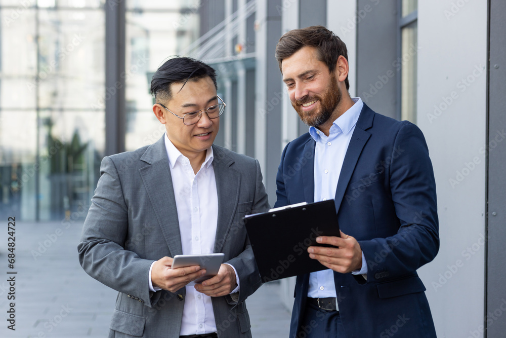 Two interracial business men standing outside an office building with a tablet and documents in their hands and discussing a plan, project, deal.