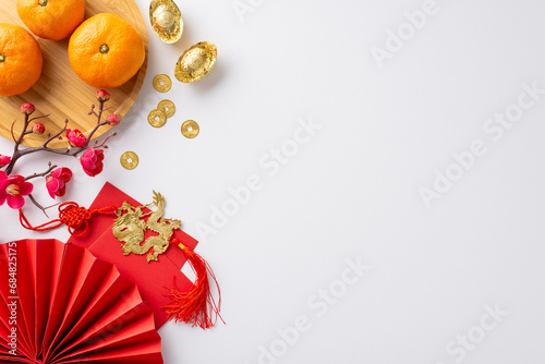Chinese New Year festivity concept. Top view of traditional coins, decorative fan, plate with mandarin oranges, golden sycee, intricate paper cutouts on festive white backdrop with space for greetings