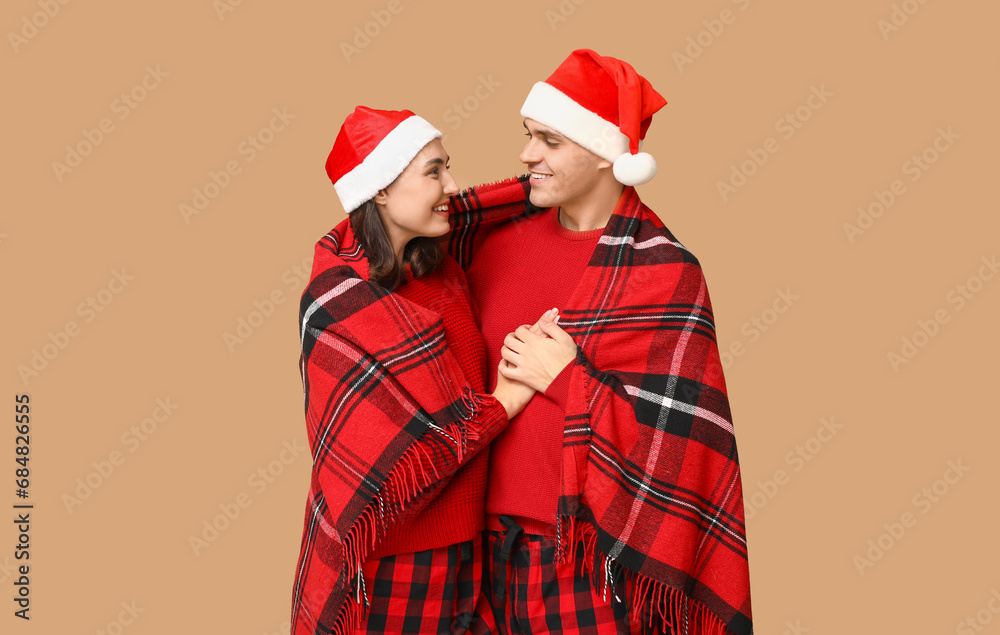 Happy young couple in Christmas pajamas and with warm plaid hugging on beige background Happy young couple in Christmas pajamas and with warm plaid hugging on beige background
