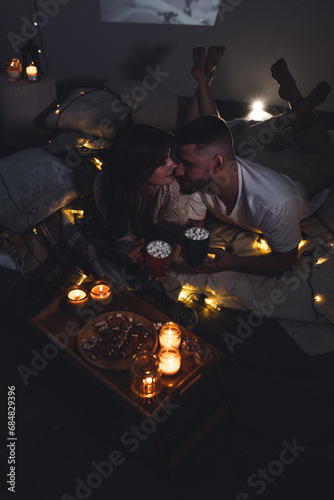 Cozy romantic evening, home weekend In bed for a loving couple. Candles, movies, hot chocolate with marshmallow, popcorn. Beautiful young man and woman enjoying relationship, having fun, laughing