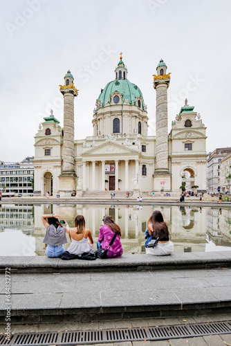 Vienna, Austria. Facade of the St. Charles Church (Karlskirche). In the foreground, a group of young people sitting at the edge of the fountain in which the church is reflected.