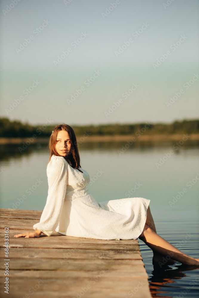 a girl in a white dress sits on a bridge on a lake in sunset in summer