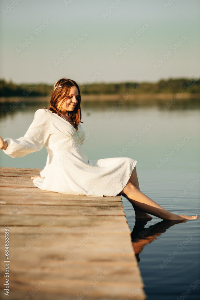 a girl in a white dress sits on a bridge on a lake in sunset in summer