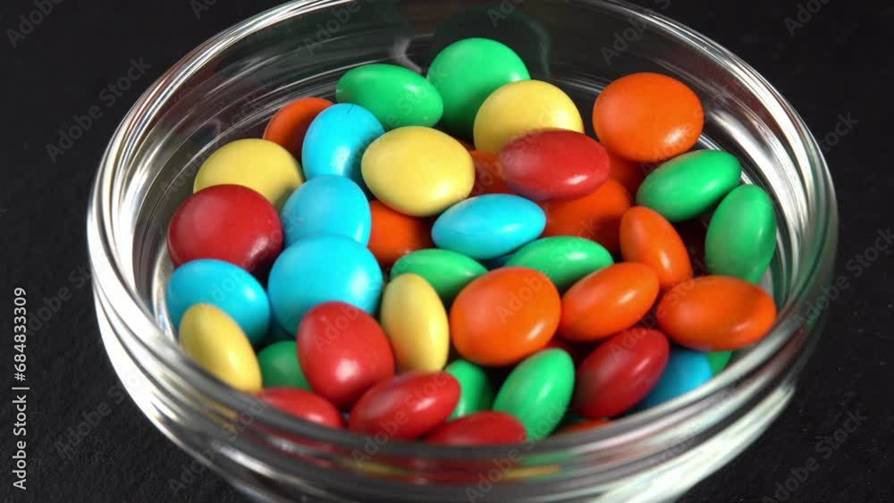 Sweet dragees in colored glaze are rotating in a glass bowl. Delicious sweets and candies close-up that children like. Chocolate Coating Sweet dragees in colored glaze are rotating in a glass bowl. Delicious sweets and candies close-up that children like. Chocolate Coating