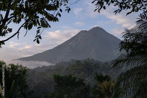 Stunning view of the Arenal volcano in the morning surrounded by the fog and tropical trees on the foreground.
