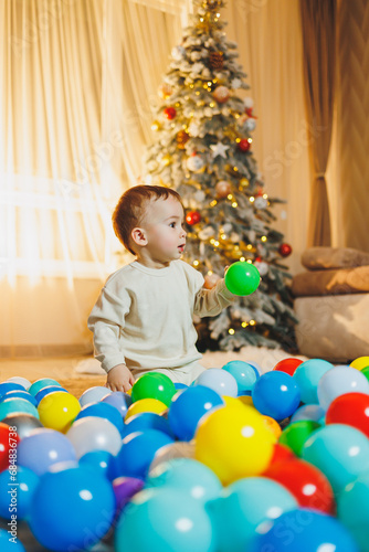 Cute boy 1 year old near the Christmas tree, sitting on the floor under the tree in the room. Holiday season. New Year. Christmas.