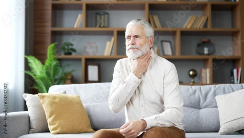 A sick elderly senior gray haired man suffering from a sore throat sitting on sofa in living room at home. Mature retired male has symptoms of a cold, virus or flu. Pensioner massaging a sore spot photo