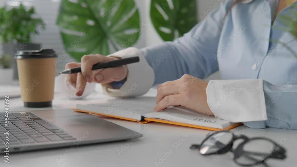 A student studies online using a computer to access materials and lessons. Woman on distance learning. Close-up of hand writing in notebook on writing desk