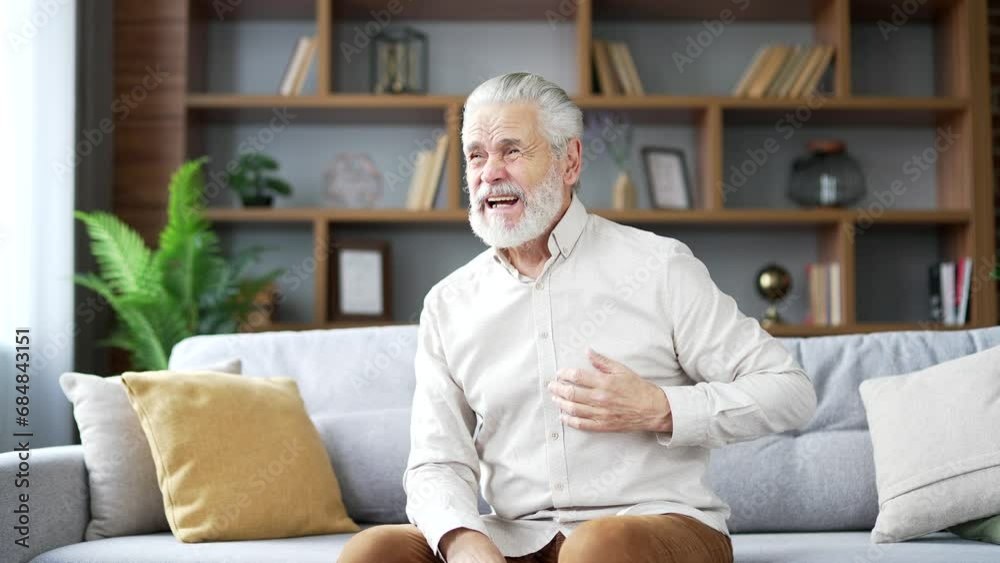 Sick elderly senior gray haired man suffering from chest tension heart attack sitting on sofa in living room at home. Adult mature male holding his chest with his hands, needs urgent medical attention Sick elderly senior gray haired man suffering from chest tension heart attack sitting on sofa in living room at home. Adult mature male holding his chest with his hands, needs urgent medical attention