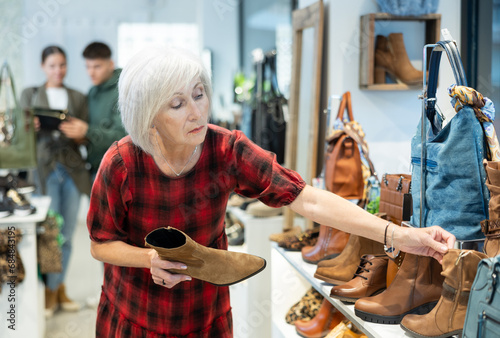 Senior woman shopper in leather product store finds it difficult to choose and thoughtfully view leather shoe boots near showcase with winter shoes. Smiling attractive woman shopping at shoes store photo