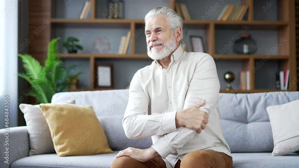 Elderly senior gray haired man suffering from elbow pain sitting on sofa in living room at home. Upset mature retired male is massaging a joint, rubs his hand. He has a joint injury or ligament sprain Elderly senior gray haired man suffering from elbow pain sitting on sofa in living room at home. Upset mature retired male is massaging a joint, rubs his hand. He has a joint injury or ligament sprain