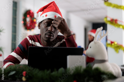 Depressed exhausted african american employee struggling with demanding company project during Christmas holiday season. Unhappy stressed worker getting overwhelmed by tasks in festive ornate office photo