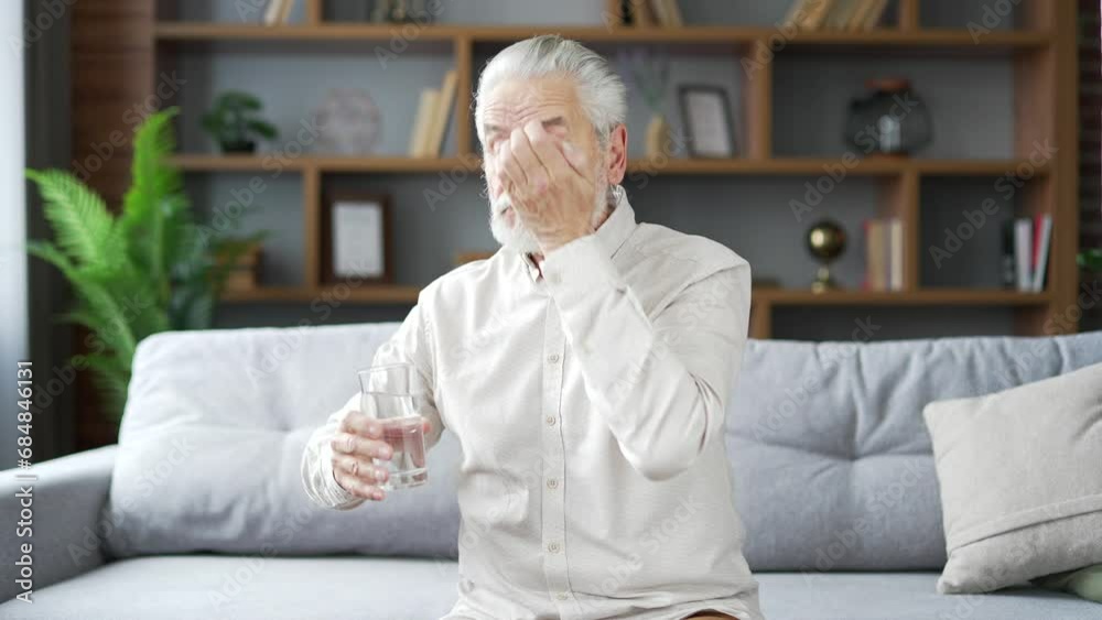 Sick elderly senior gray haired bearded man takes painkillers and drinks water from a glass while sitting on sofa in living room at home. Overtired adult mature grandfather suffering from headache Sick elderly senior gray haired bearded man takes painkillers and drinks water from a glass while sitting on sofa in living room at home. Overtired adult mature grandfather suffering from headache