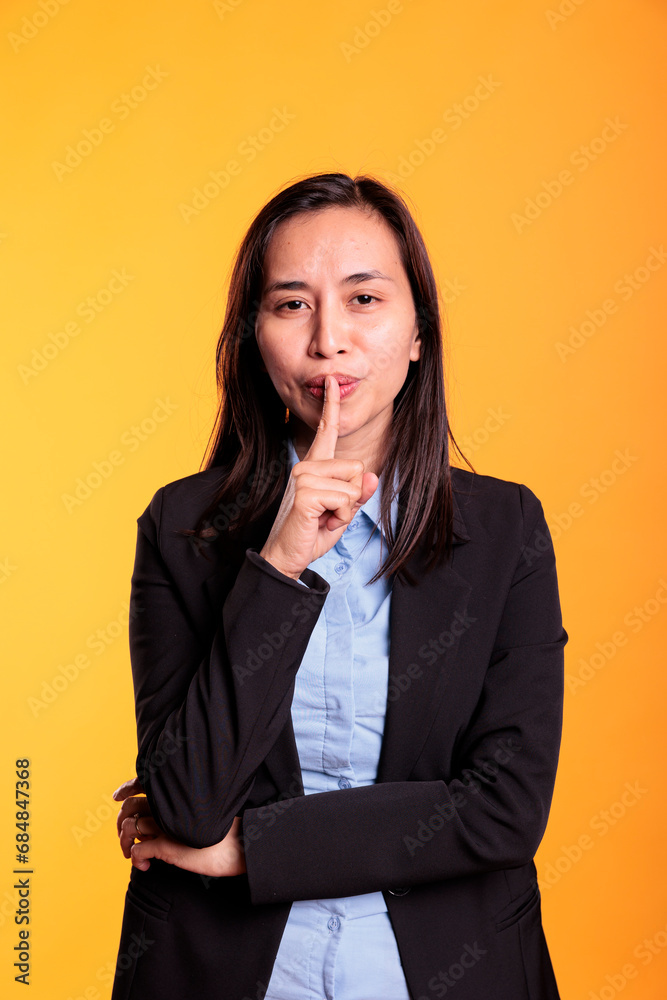 Portrait of filipino woman making shh gesture in studio by putting forefinger over lips posing over yellow background. Silent adult shushing people gesturing secrecy and confidentiality. Portrait of filipino woman making shh gesture in studio by putting forefinger over lips posing over yellow background. Silent adult shushing people gesturing secrecy and confidentiality.