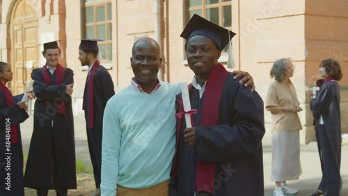Portrait of young African American man in gown and hat posing for photo together with his father against university photo