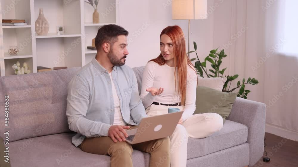 Happy smiling young family couple talking sitting on the couch with laptop computer browsing product in online internet store planning apartment decoration and imagine their beautiful house together Happy smiling young family couple talking sitting on the couch with laptop computer browsing product in online internet store planning apartment decoration and imagine their beautiful house together