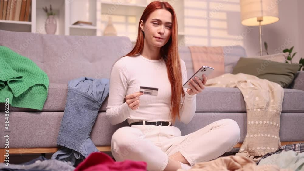 Pretty young woman paying with credit card on smartphone while sitting on clothes on the floor Happy customer doing payments online shopping in internet store because there is nothing to wear at home Pretty young woman paying with credit card on smartphone while sitting on clothes on the floor Happy customer doing payments online shopping in internet store because there is nothing to wear at home