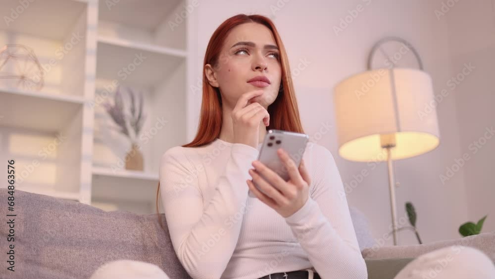Portrait of creative red haired woman student hold smartphone and thinks questioningly about difficult choice choose imagine plan in mind has doubts create idea sitting on couch at home Portrait of creative red haired woman student hold smartphone and thinks questioningly about difficult choice choose imagine plan in mind has doubts create idea sitting on couch at home