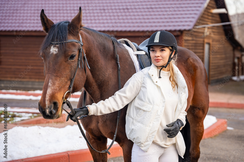 Blond professional female jockey standing near brown horse on farm in winter. Friendship with horse concept Blond professional female jockey standing near brown horse on farm in winter. Friendship with horse concept