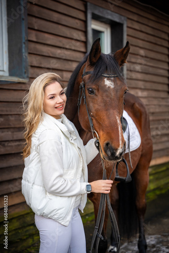 Young beautiful rider woman blonde with long hair in white clothes standing near brown horse on farm