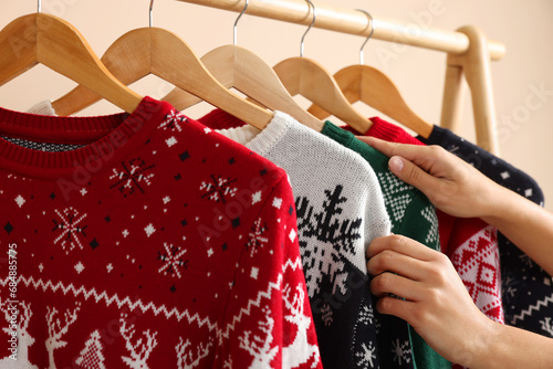 Woman choosing Christmas sweater from rack near beige wall, closeup