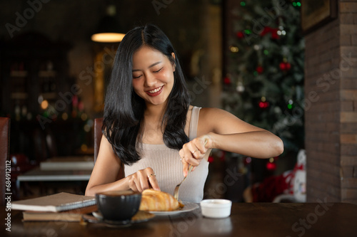 Elegant young Asian woman eating croissant fro breakfast with hot latte coffee in a dark luxury living room.