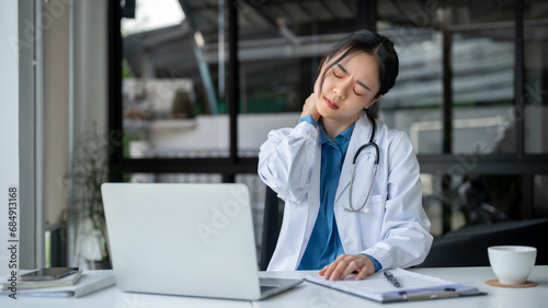 A tired doctor is massaging her neck, suffering from neck ache and office syndrome while working.