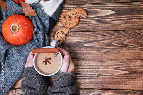 Pretty female hands warming fresh coffee mug, latte, spicy cortado, bright orange pumpkin wrapped in woolen scarf, sweet biscuits, dry oak leaves, spices on rustic wooden table. Autumn background