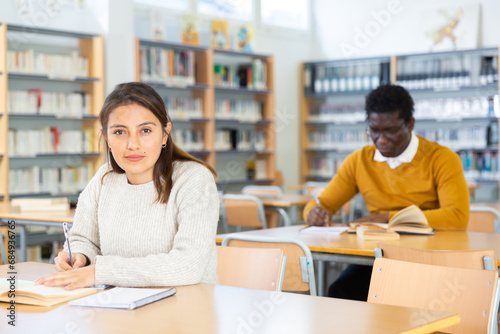Portrait of positive young hispanic woman sitting at table in public library. Adult self-learning concept