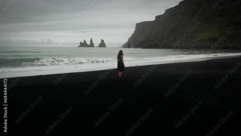 Young beautiful woman in black dress walking on black sand beach, epic aerial ocean scenery, Iceland
