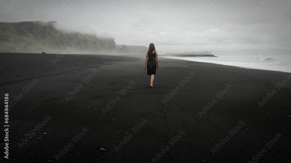 Young beautiful woman in black dress walking on black sand beach Iceland, misty dramatic mountain landscape, tracking shot
