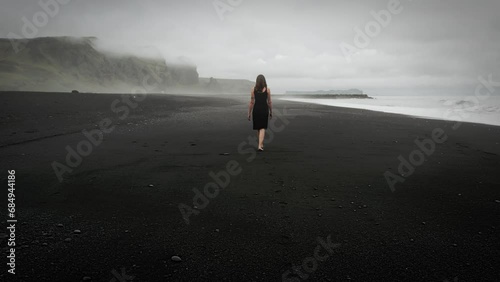 Young beautiful woman in black dress walking on black sand beach Iceland, misty dramatic mountain landscape, tracking shot photo