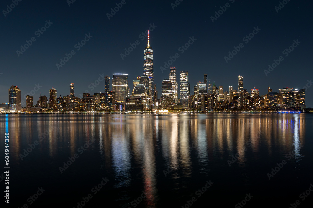 New York City's Manhattan skyline at night, photographed from Jersey City with Hudson River in front.