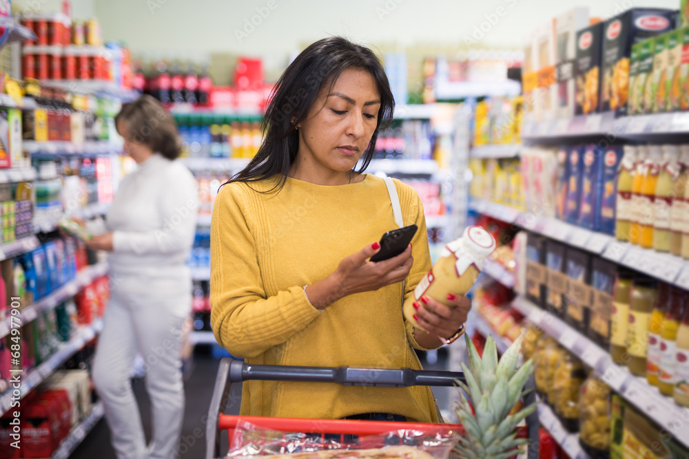 Latin american woman making purchases in grocery store, using smartphone to scan barcode of products. Modern shopping concept