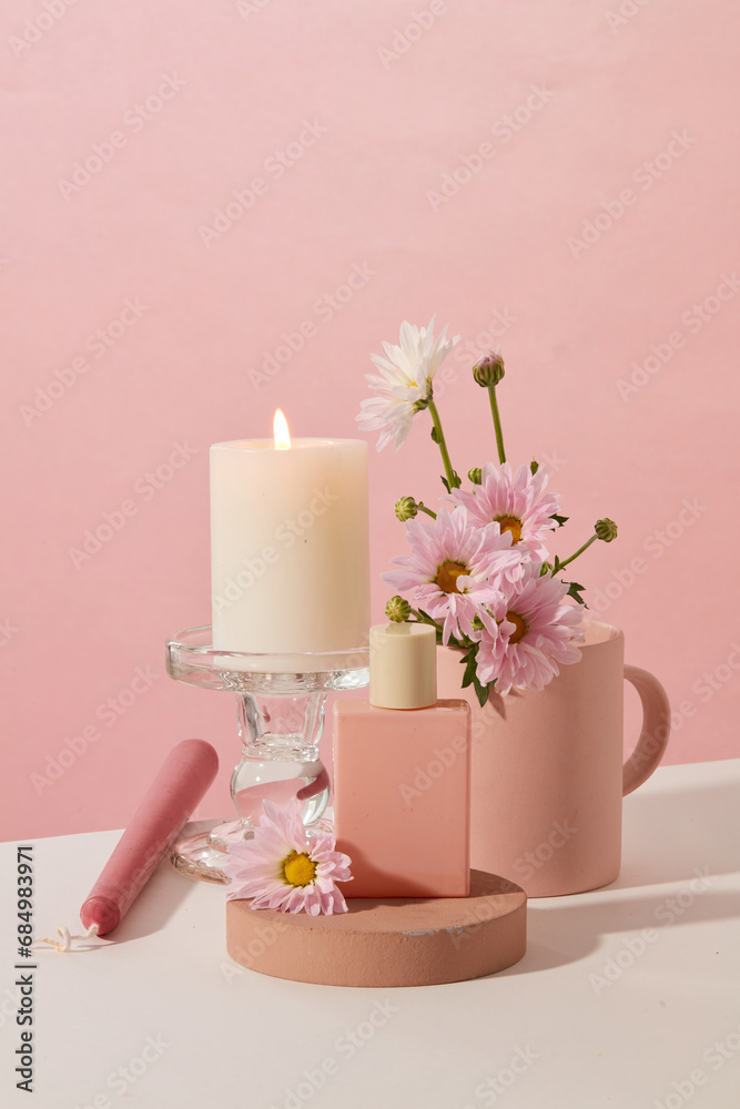 A burning candle is placed on a glass candlestick, an unlabeled perfume bottle is placed on a wooden podium and fresh flowers are on a pink and white background. Copy space for ads.