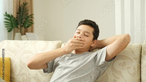 Tired sad teen boy in a gray t shirt sitting on the couch to watch television. Bored teenager with hands on head yawns.