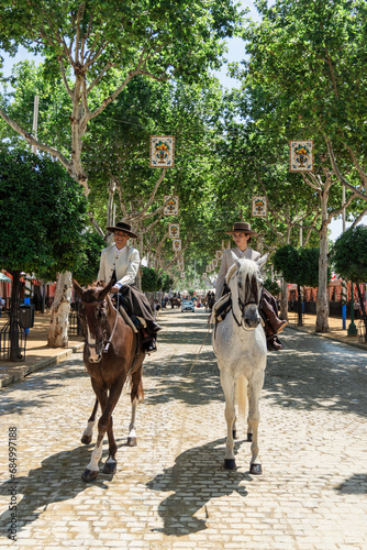 Young diverse women in hats riding horses under tree shade during daytime