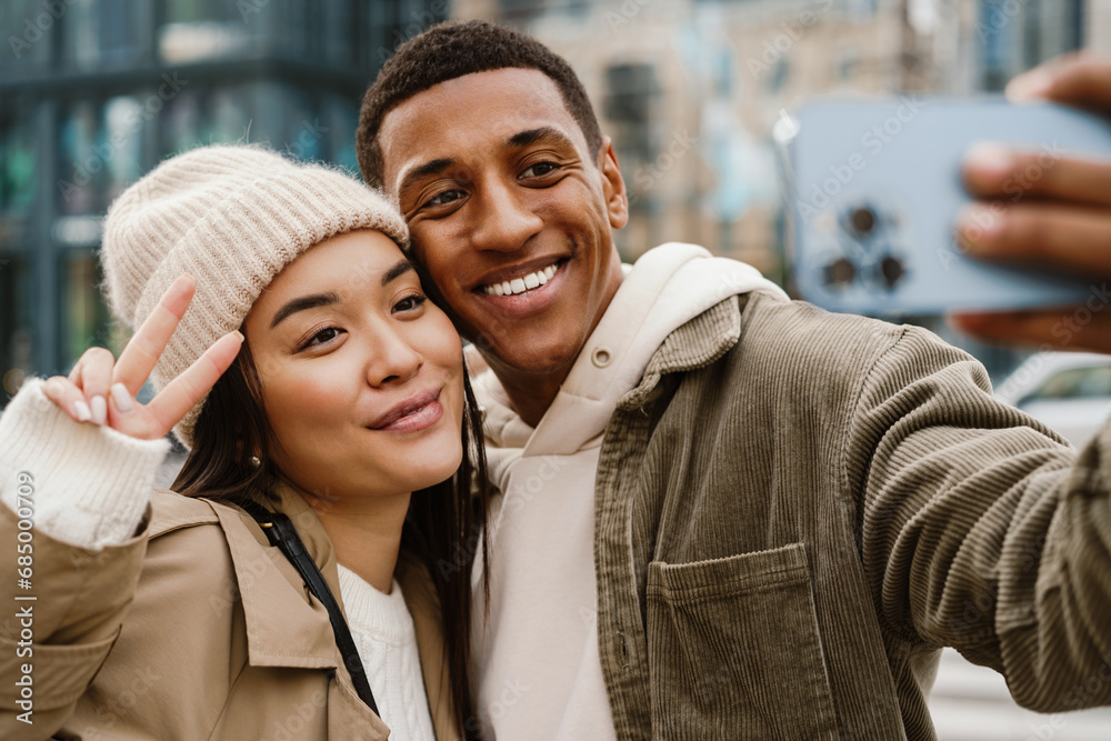 Smiling couple taking selfies with smartphone camera and showing peace sign while standing at street
