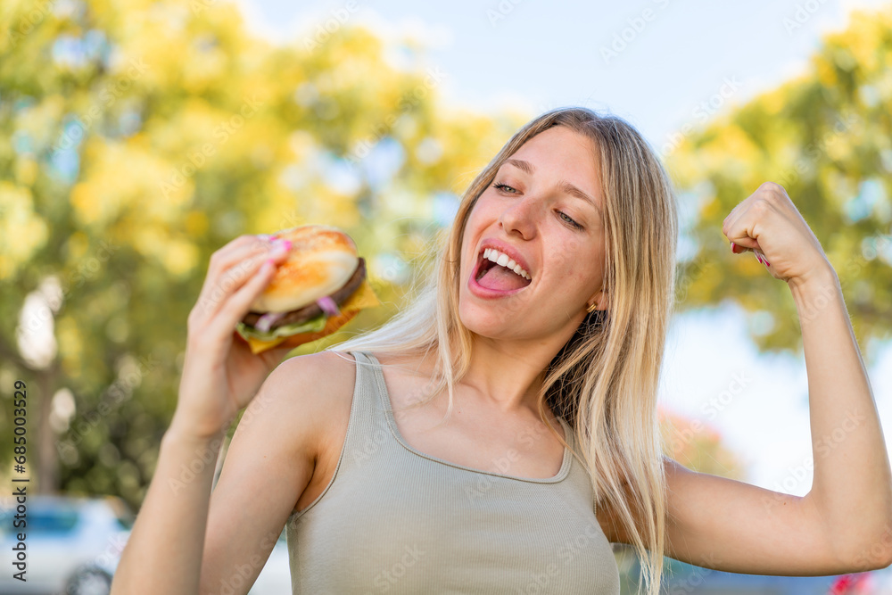 Young blonde woman holding a burger at outdoors celebrating a victory