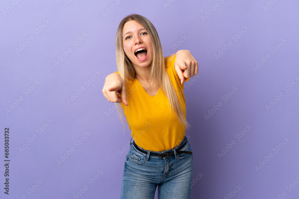 Young blonde woman isolated on purple background surprised and pointing front Young blonde woman isolated on purple background surprised and pointing front