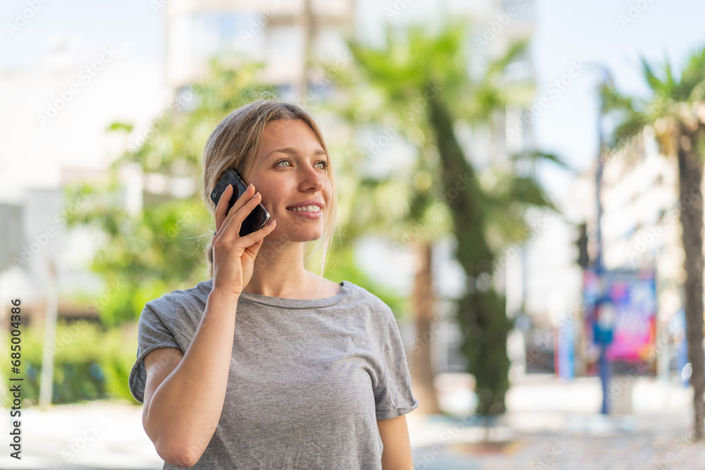 Young blonde woman at outdoors keeping a conversation with the mobile phone with someone Young blonde woman at outdoors keeping a conversation with the mobile phone with someone