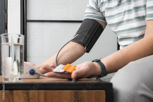 Unrecognizable man sitting on a sofa at home and measures blood pressure on a hand with modern digital tonometer and holding blister packs with pills, medicines