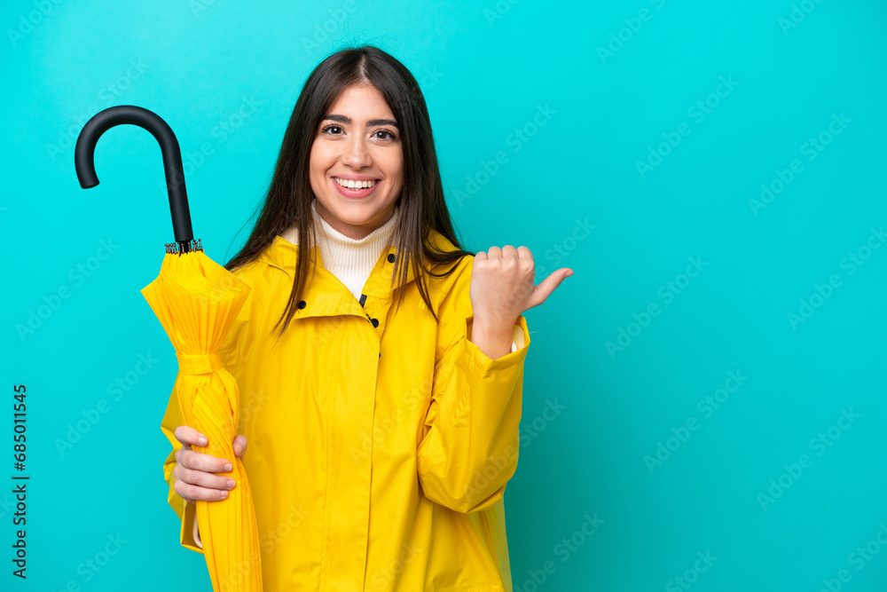 Young caucasian woman with rainproof coat and umbrella isolated on blue background pointing to the side to present a product Young caucasian woman with rainproof coat and umbrella isolated on blue background pointing to the side to present a product