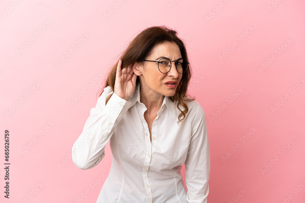 Middle aged caucasian woman isolated on pink background listening to something by putting hand on the ear