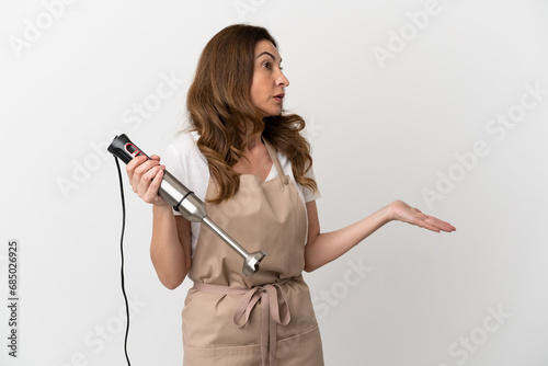 Middle aged caucasian woman using hand blender isolated on white background with surprise expression while looking side